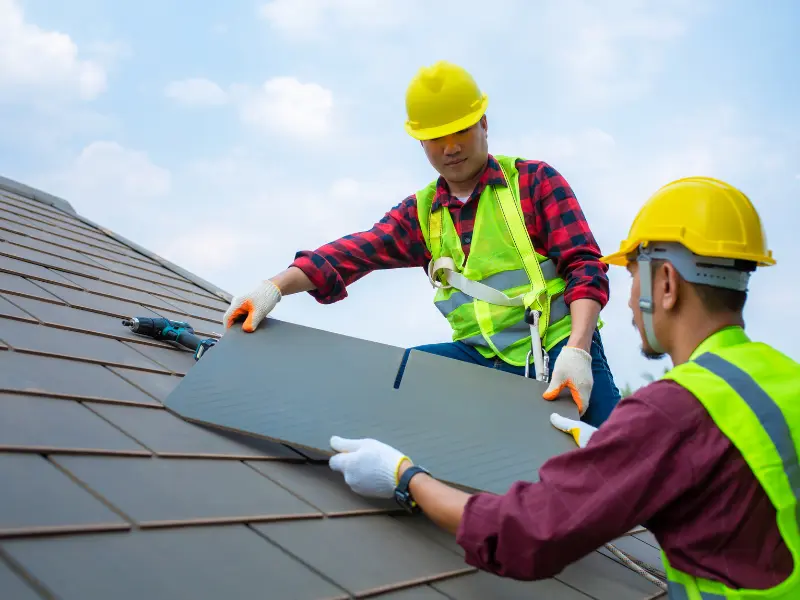 Two roofers in hard hats installing a gray roof tile.