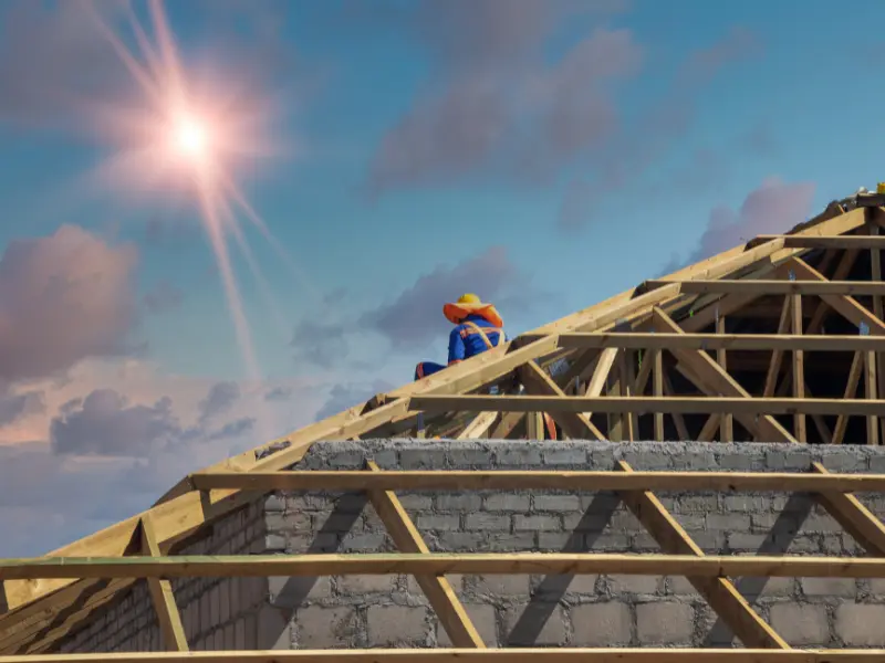 Worker sitting atop wooden roof trusses under a bright sun.